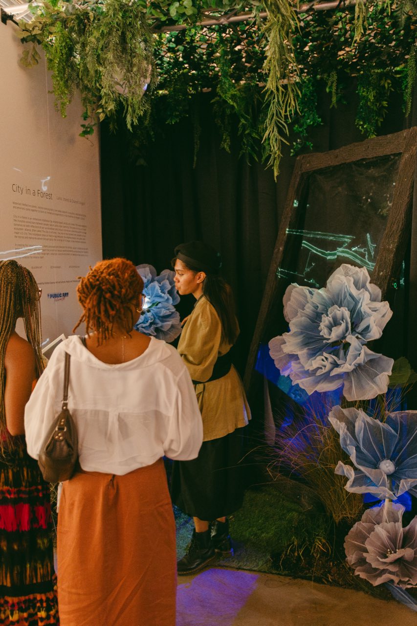 Visitors gather as the artist points to a glowing blue projection on the Pepper's Ghost panel, greenery hanging overhead.