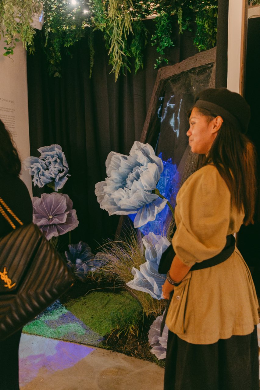 A visitor in gold pausing before the towering blue paper flowers.